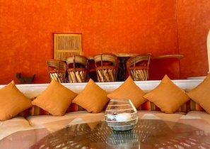 Un coin salon avec une table en verre, des coussins orange et des chaises en osier, le tout adossé à un mur rouge.