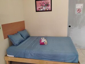 Ein Einzelbett mit Holz-Kopfteil, einer blauen Matratzenauflage und zwei Kissen. Ein kleines, pinkfarbenes Accessoire liegt auf dem Bett.