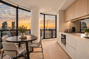 A modern kitchen with a city view, a dining area with a table and chairs, and a large window overlooking the sunset. - Sub Penthouse in Docklands with Breathtaking 180° Views | Pool, Gym & Parking (Docklands)