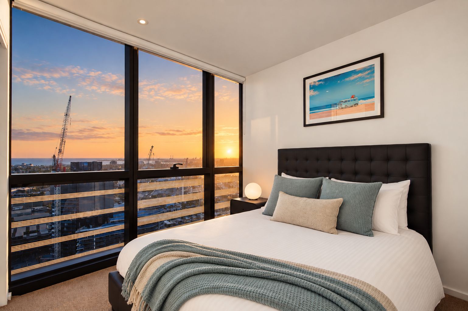 A bedroom with a large bed, a nightstand, a framed picture, and a view of a sunset over a cityscape.