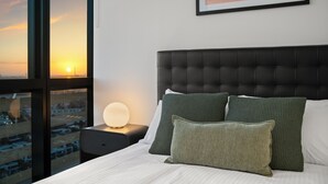Hotellihuone, jossa on suuri ikkuna, vihreä- ja valkoisilla tekstiileillä varustettu sänky, musta pääty ja kehystetty kuva rantamaisemasta.