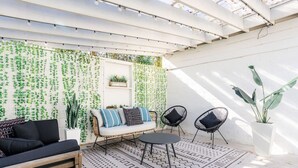 Un patio cubierto con una pared blanca, plantas colgantes, un sofá, dos sillones y una mesa.