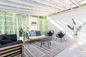 Un patio cubierto con una pared blanca, plantas colgantes, un sofá, dos sillones y una mesa.