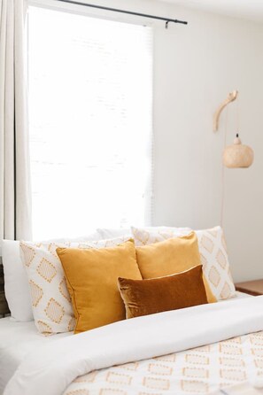 Una cama con almohadas estampadas, una lámpara colgante y un ventanal con cortinas.