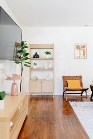 Een kamer met een houten vloer, een houten stoel met een geel kussen, een houten kast met planken, een potplant en een lijst met een kunstwerk aan de muur.