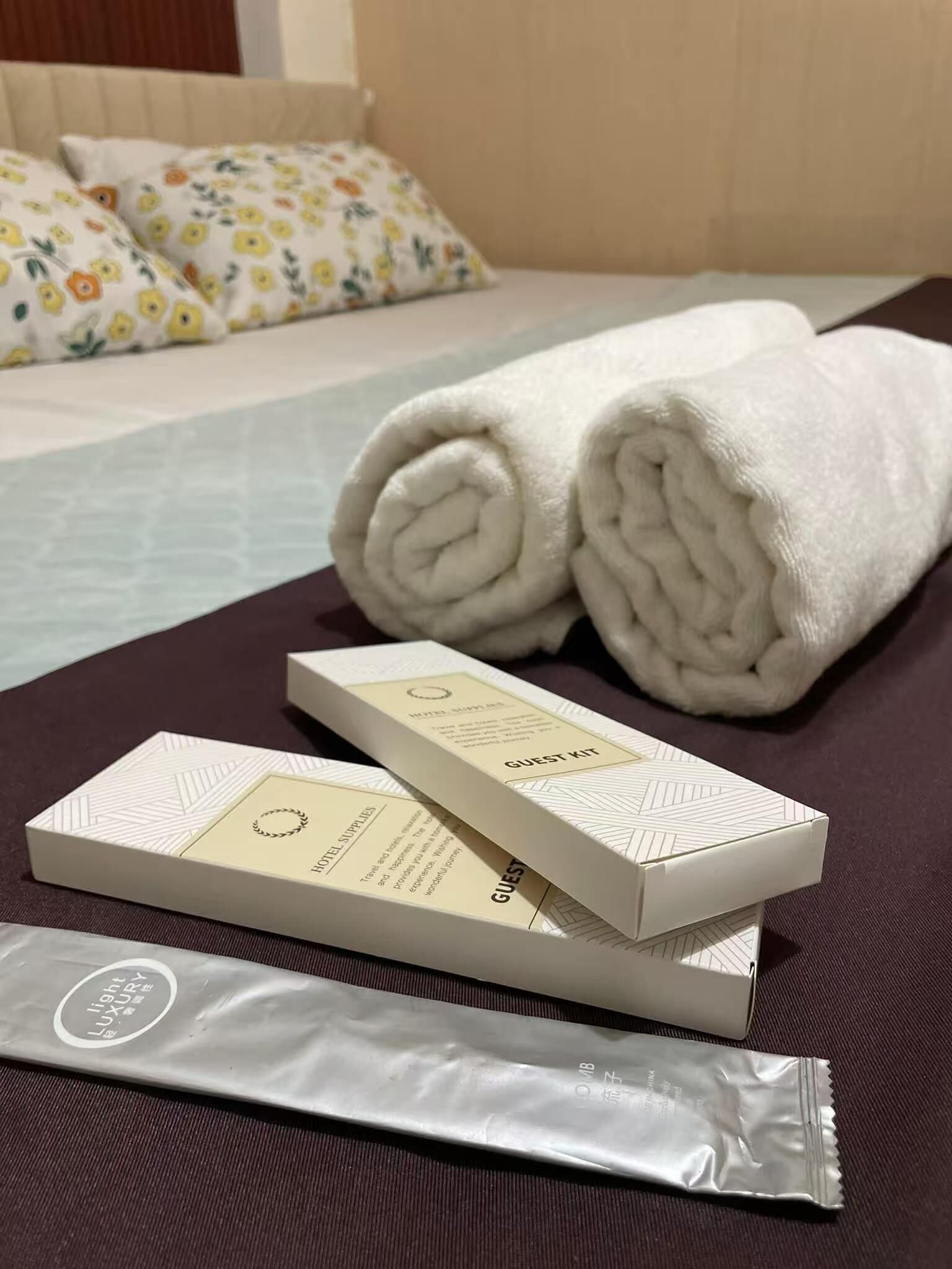 Un lit bien fait, agrémenté de serviettes blanches pliées, d’une boîte contenant des articles de toilette pour les invités et d’un produit de soin pour la peau, le tout posé sur une surface en tissu foncé.