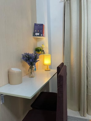 Un coin de pièce avec un bureau, une lampe, une poubelle, une petite plante et une bibliothèque.
