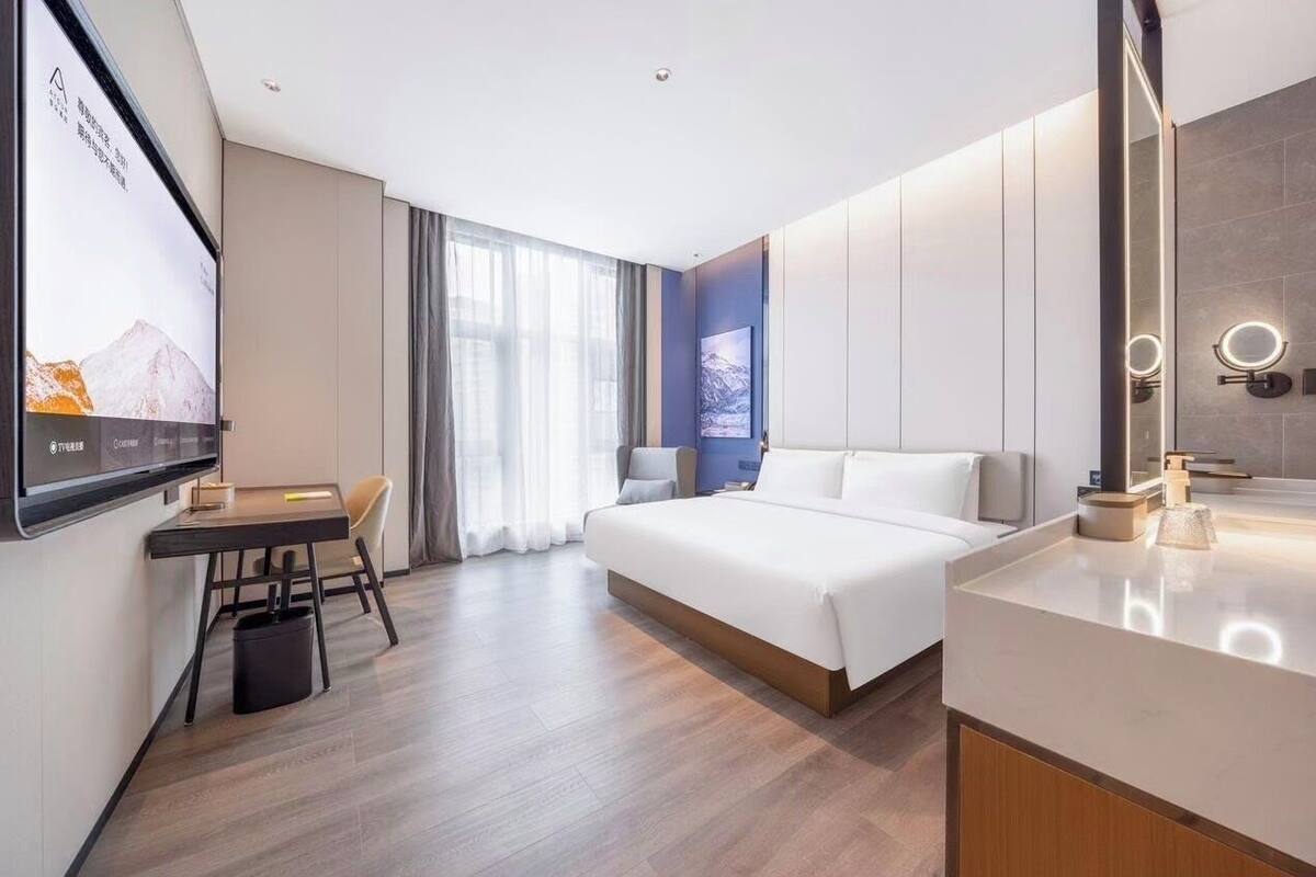 A modern hotel room with a bed, a desk, a chair, a TV, and a bathroom with a mirror and lights.