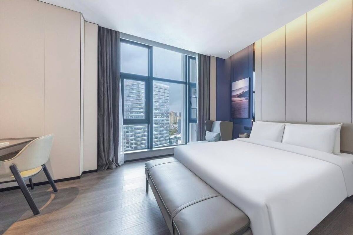 A modern hotel room with a large bed, a desk, and a view of the cityscape through floor-to-ceiling windows.