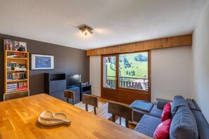 Ein Zimmer mit einem Holzespeisetisch, Stühlen, einer Couch, einem Fernseher, einem Bücherregal und einem großen Fenster mit Blick auf eine grüne Landschaft.