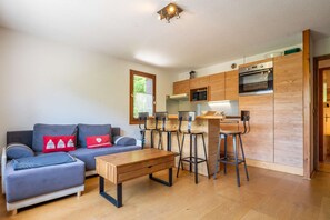 Ein kompakter Wohnbereich mit einer Couch, einem Holzesstisch, Barhocker, einer Küchenzeile mit Mikrowelle und Backofen sowie einem Fenster mit Blick.