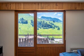 A room with a large window offering a view of a green landscape and a building. - Gerfaut : Centre à pied, vue montagnes (LE GRAND BORNAND)