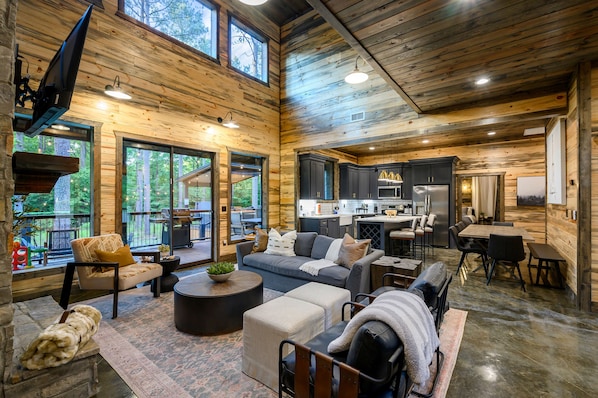 A spacious living area with a sofa, coffee table, and dining table, connected to a kitchen with modern appliances. - New Cabin/Hot Tub/Fire Pit/Arcade/Shuffleboard (Broken Bow)