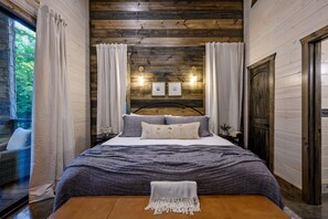 Een slaapkamer met een houten hoofdbord, een bed met grijze beddengoed, twee wandlampen en een raam met gordijnen.