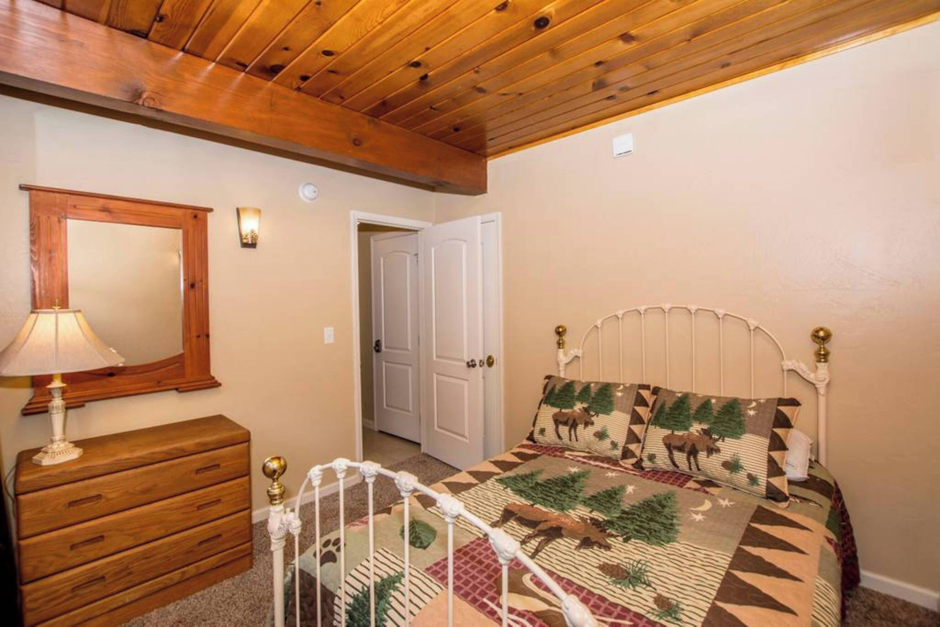 A bedroom with a wooden ceiling, a bed with a patterned bedspread, a dresser, a mirror, and a door.
