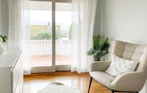 Ein Zimmer mit einer Schiebeglastür, die auf einen Balkon führt, einem beigen Sessel, einem weißen Teppich und einem Blumentopf.