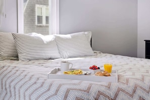 A neatly made bed with striped pillows, a tray with breakfast including toast, fruit, and juice, and a window with a view of a building.