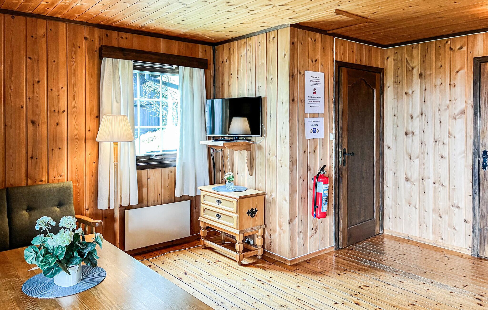 Una habitación con paneles de madera, un televisor, un mueble pequeño, un radiador, un extintor y una ventana con cortinas.