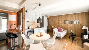 Eine moderne Küche mit weißen Schränken, einem Holzes Esstisch mit vier weißen Stühlen, einer Couch mit einem rosafarbenen Kissen und einer Holzwand mit einer Bücherreihe.