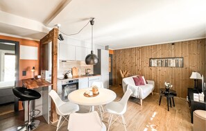 Eine moderne Küche mit weißen Schränken, einem Holzes Esstisch mit vier weißen Stühlen, einer Couch mit einem rosafarbenen Kissen und einer Holzwand mit einer Bücherreihe.
