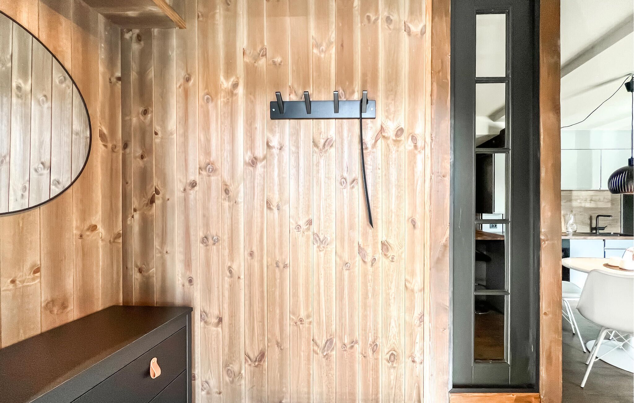 Een kamer met een houten wand, een zwart kapstokje, een spiegel en een donkere dressoir.