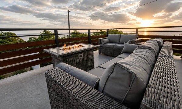 An outdoor seating area with a fire feature, wicker furniture, and a view of the sunset over the water.