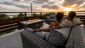 A couple relaxing on a wicker sofa with a glass-top table and a fire feature, overlooking a body of water during sunset.