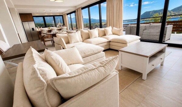 A modern living room with a large L-shaped sofa, a dining area, and a balcony with a scenic mountain view.