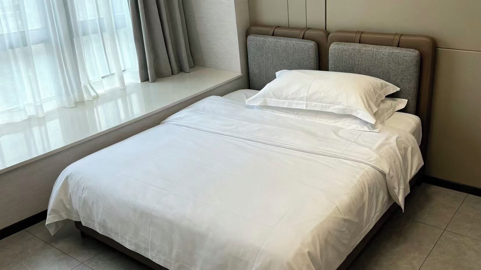 A neatly made bed with a headboard, a window with curtains, and a tiled floor.