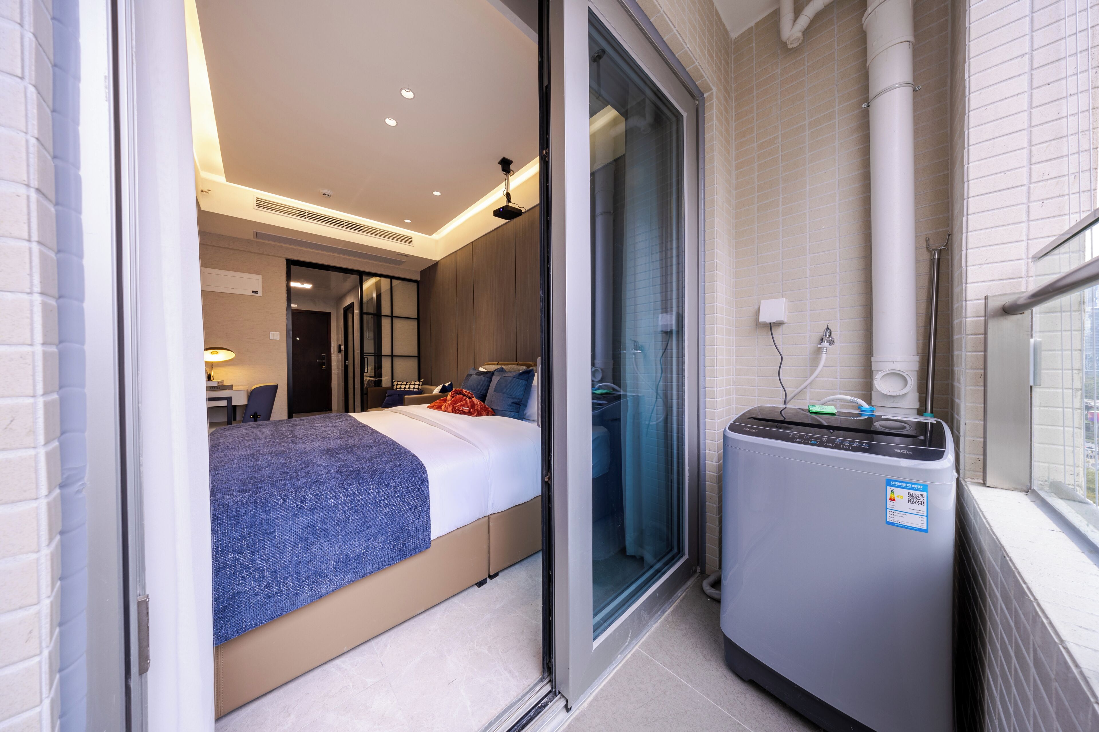 A hotel room with a bed, a small desk, a shower area, and a compact washing machine.