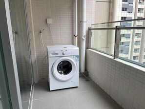 A compact, modern laundry room with a front-loading washing machine, a glass shower enclosure, and a view of a high-rise building through a large window. - BrisAudio-Visual X Ju (Shenzhen)
