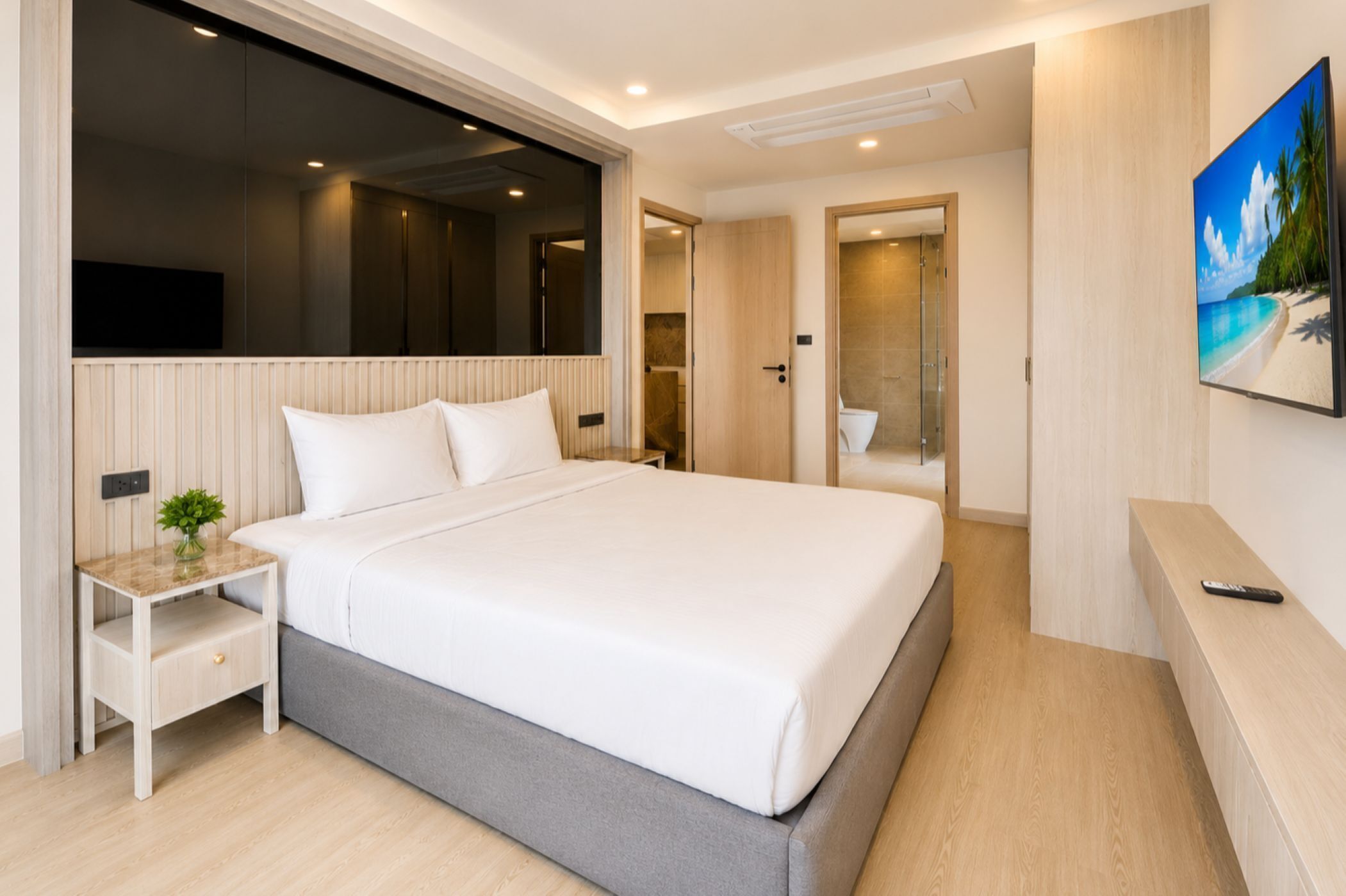 A modern hotel room with a large bed, a bedside table with a plant, a TV mounted on the wall, and a bathroom visible through an open door.
