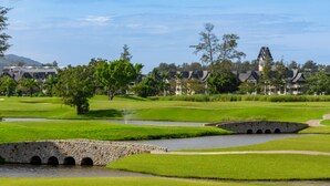 A golf course with a stone bridge, water hazards, and traditional-style buildings in the background. - New 2 BR Apt Golf View - Ozone 108-156 (Thalang)