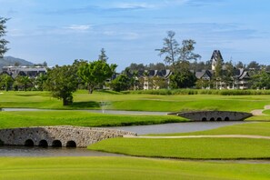Um campo de golfe com uma ponte de pedra, zonas de água e edifícios em estilo tradicional ao fundo.