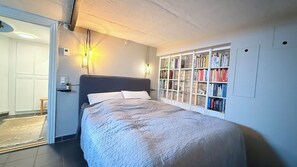 Une chambre à coucher comprenant un lit, des lampes de chevet, une bibliothèque et une porte donnant sur une autre pièce.