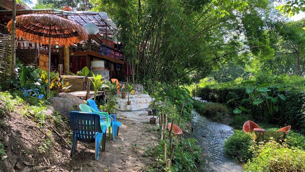 Un área rústica al aire libre con una estructura de techo de paja, un pequeño arroyo y un sendero de tierra que conduce a una escalera de madera.