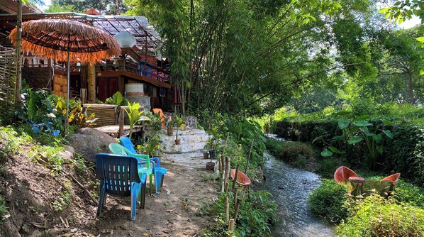 A rustic outdoor area with a thatched roof structure, a small stream, and a dirt path leading to a wooden staircase.