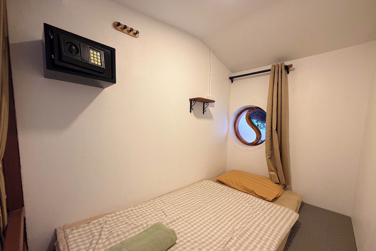 A small, single-bed room with a checkered bedspread, a green towel, a wall-mounted radio, a small shelf, and a window with a curtain.