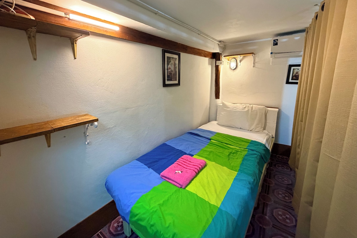 A small, single-bed room with a colorful bedspread, a wooden shelf, and a wall-mounted light fixture.