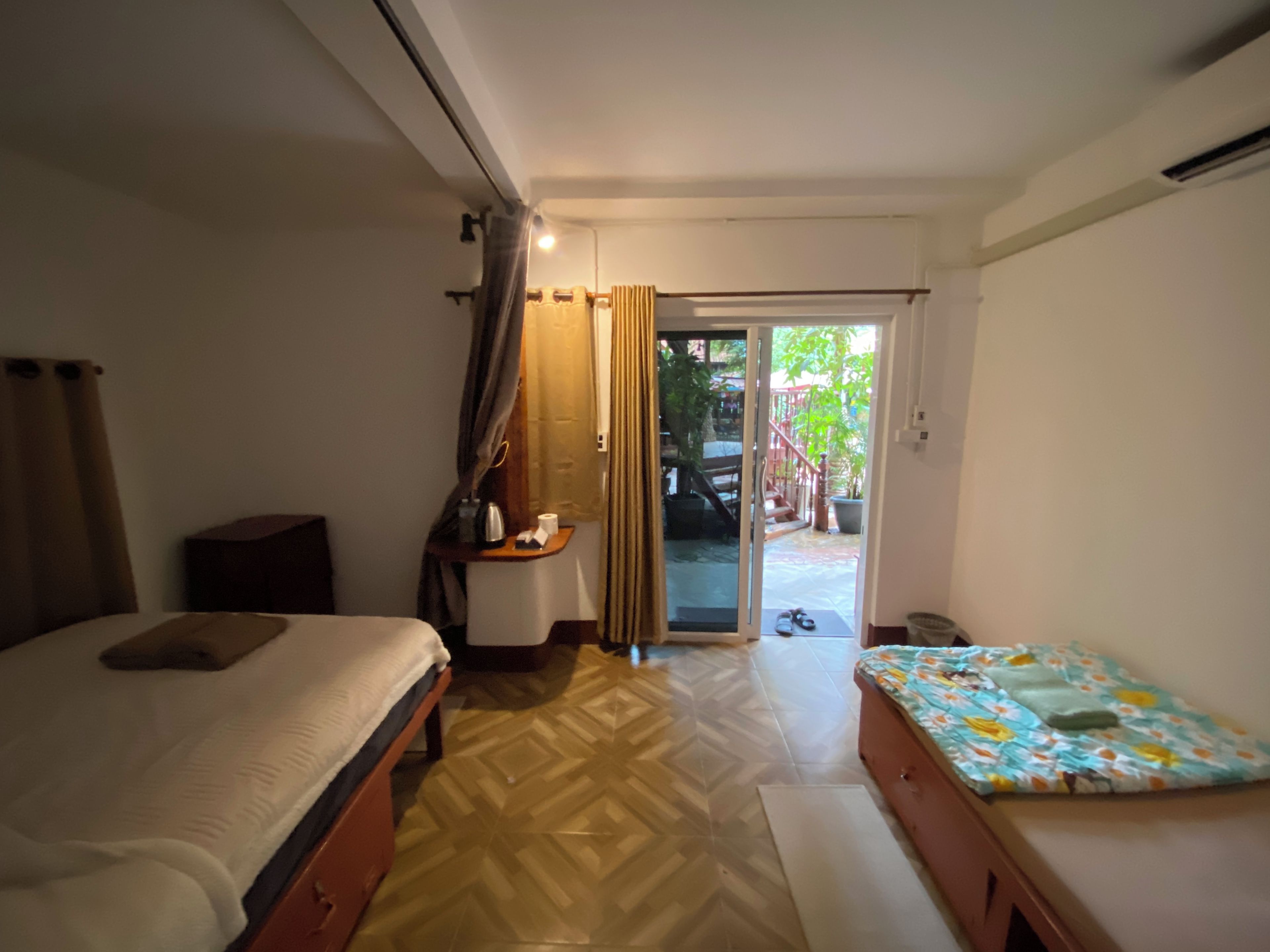 Habitación de hotel con cama, una mesita con lámpara, un banco y un ventanal corredero que da a un área exterior con escaleras y vegetación.