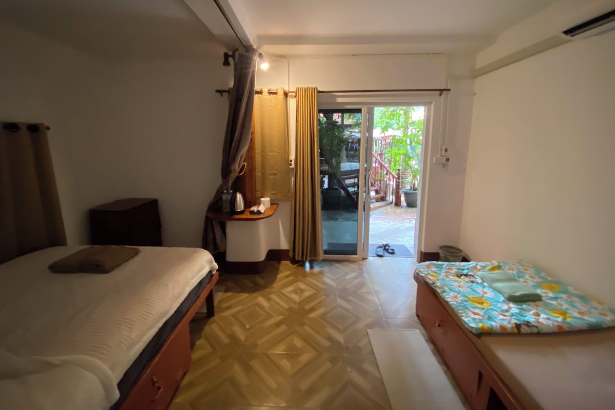 A hotel room with a bed, a small table with a lamp, a bench, and a sliding glass door leading to an outdoor area with a staircase and greenery.