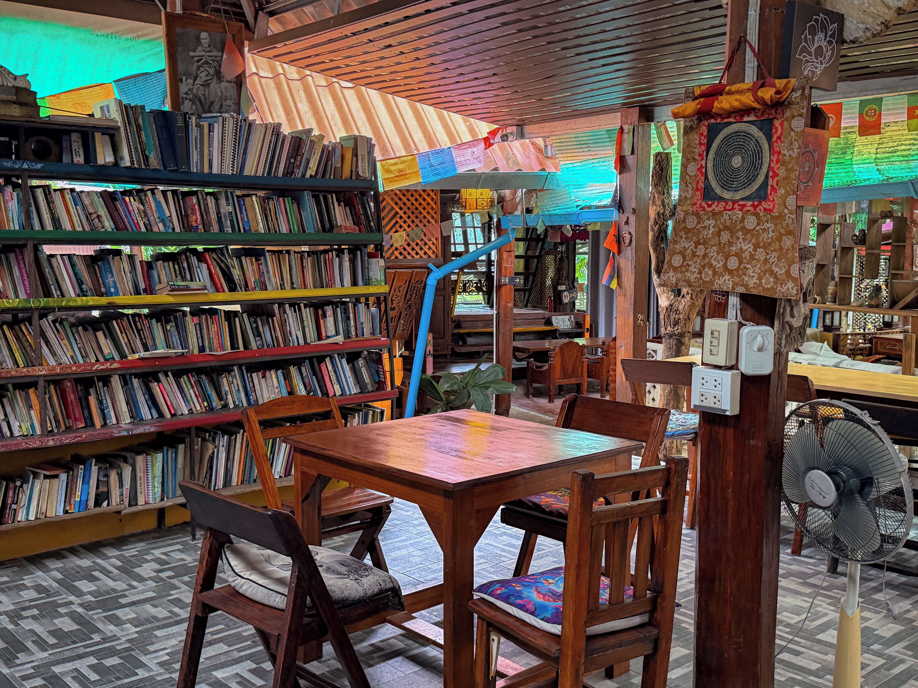 A library with multiple wooden bookshelves filled with books, a wooden table with chairs, and a fan.