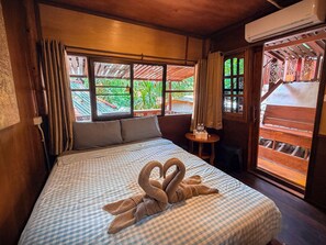 A hotel room with a bed, a small table with a chair, a window with curtains, and an air conditioning unit.