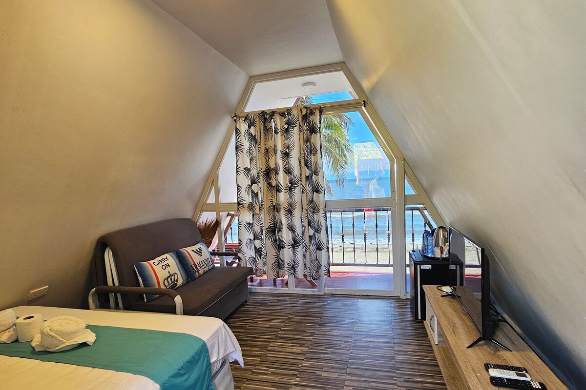 A modern hotel room with a sloped ceiling, a bed with a teal bedspread, a wooden bench, a desk with a computer, and a view of the beach through a large window.
