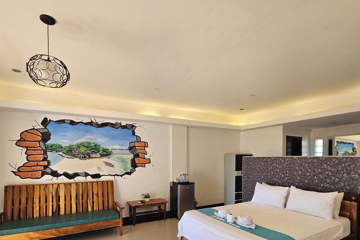 A hotel room with a bed, a wooden bench, a small table, and a wall mural.
