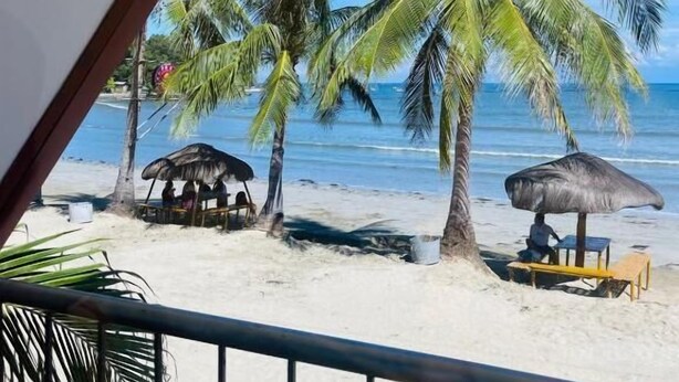 Vista de playa con palmeras, una cabaña de techo de paja y una mesa de madera con sombrilla.