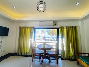 A room with a round table and chairs, a television mounted on the wall, an air conditioning unit, and a view of the beach through large windows with yellow curtains. - Bolo Hills Beach Club (Alaminos)