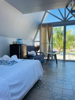 A hotel room with a bed, a desk, a chair, a small table, and a view of palm trees through large windows. - Bolo Hills Beach Club (Alaminos)