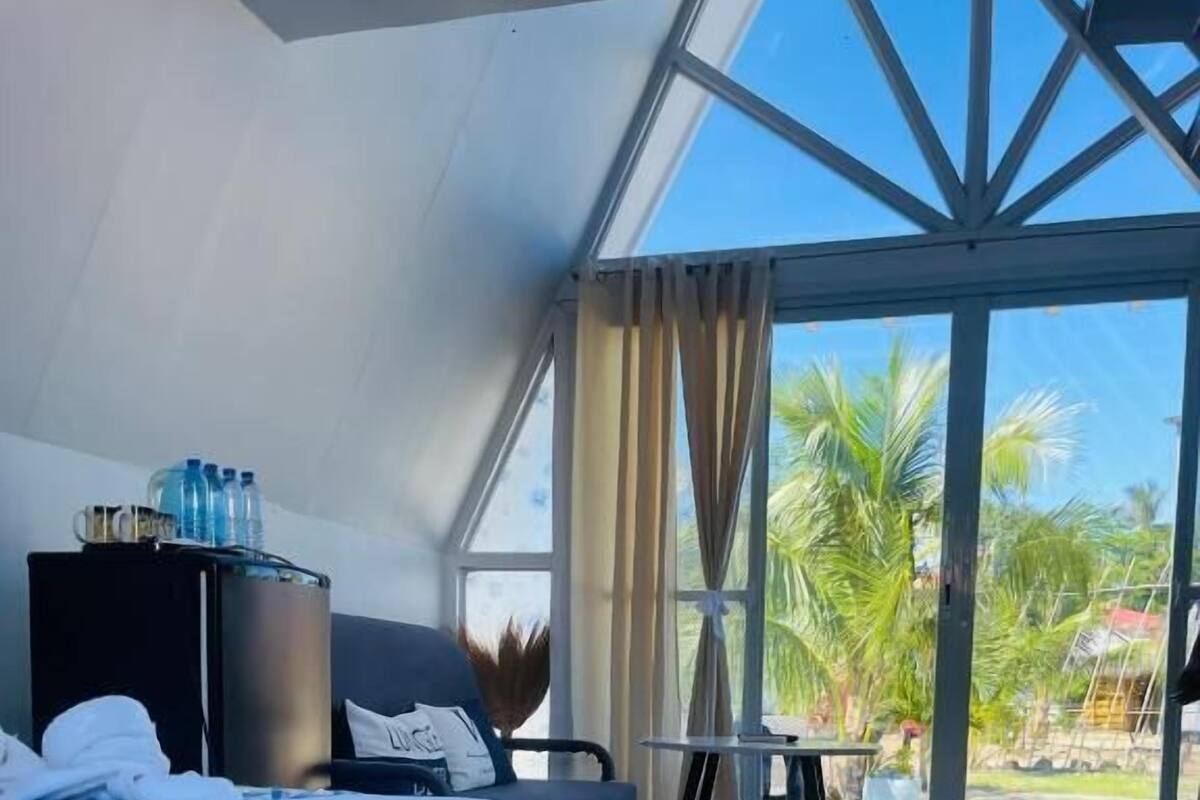 A hotel room with a bed, a small table, a chair, a water dispenser, and a view of palm trees through large windows.