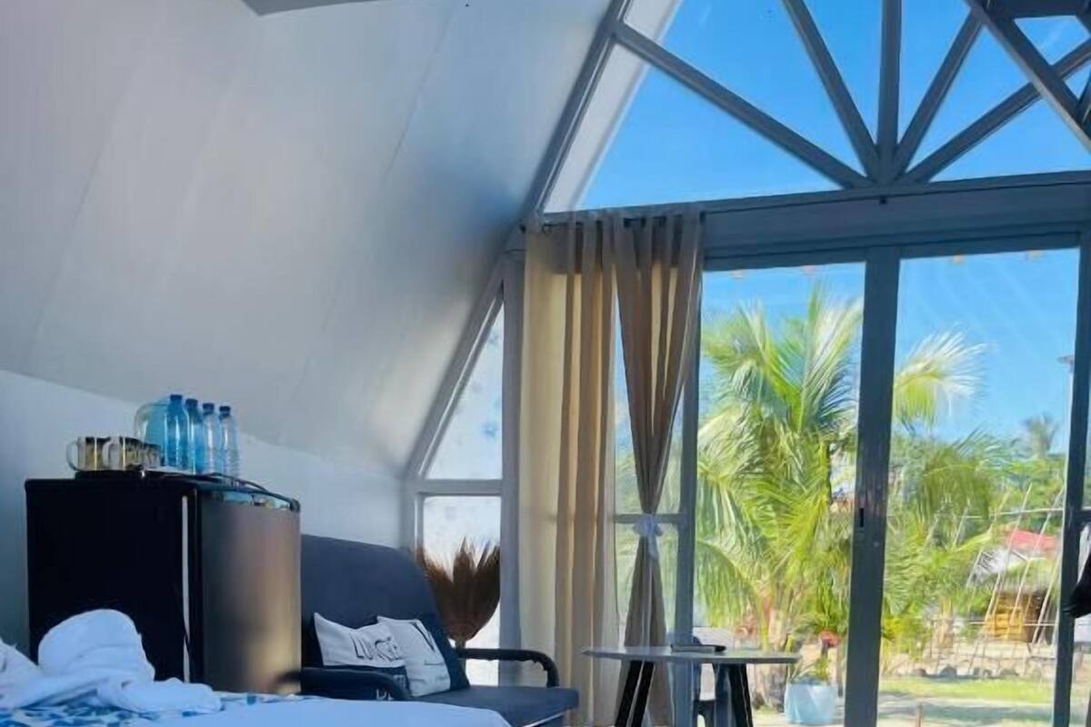 A hotel room with a bed, a small table, a chair, a minibar, and a view of palm trees through large glass doors.